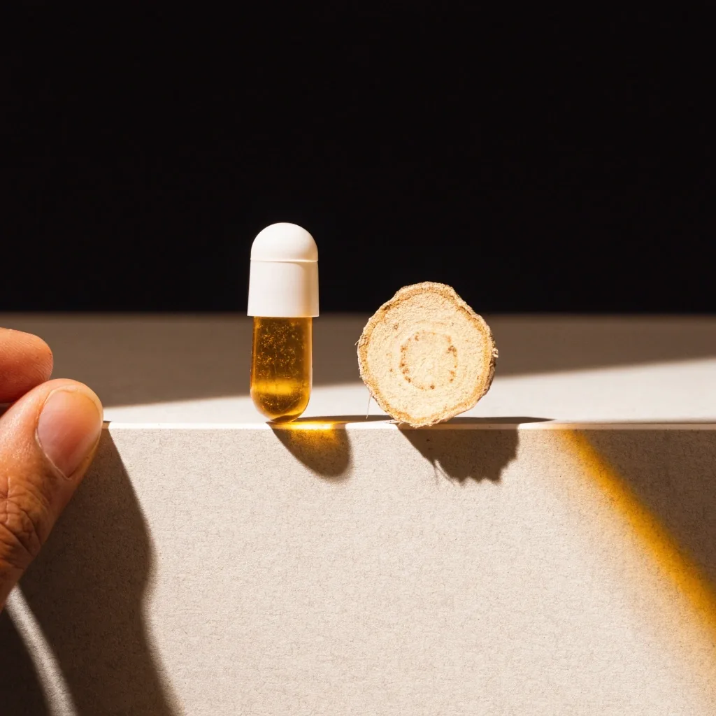 A softgel capsule and a root slice set by hand on a matte counter under a single warm bulb